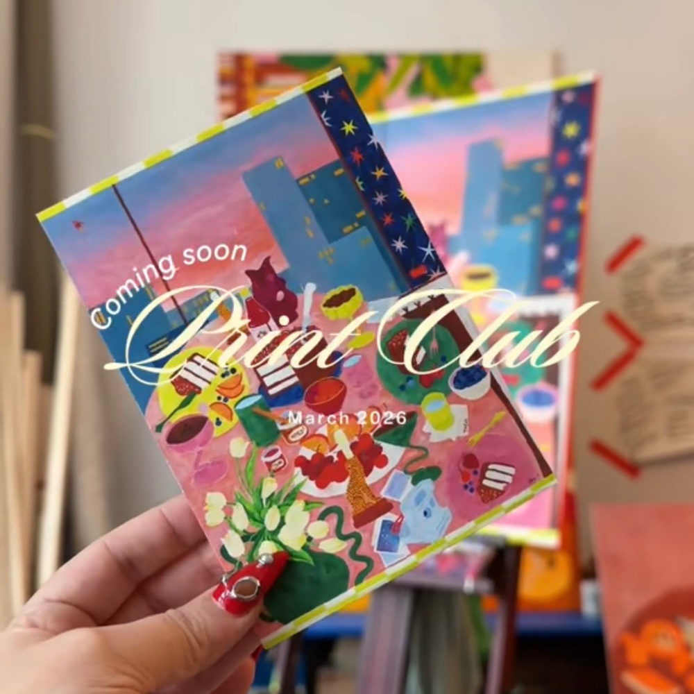 'Print Club Coming Soon' written on the top of the image and a tiny colourful art print held by a hand with a blurred background of the artist's studio. 